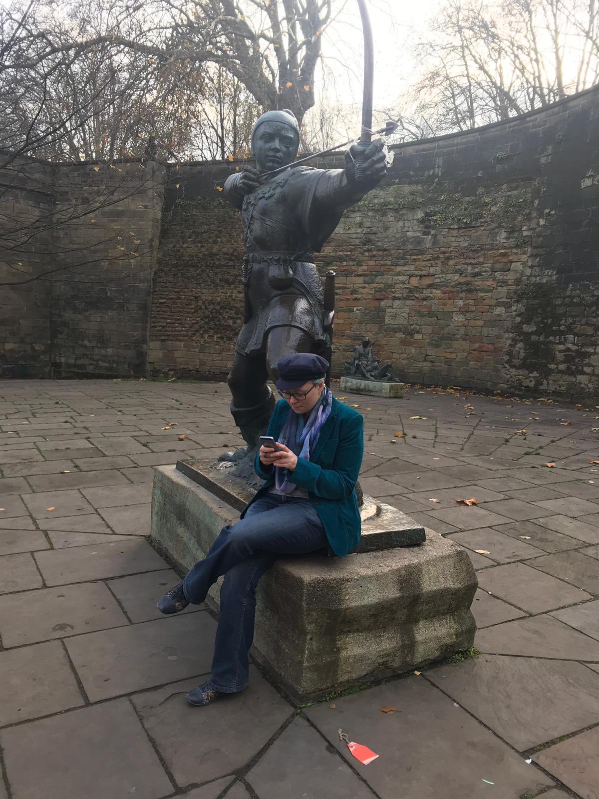 A person in a green jacket and blue hat sits on the pedestal of a statue of Robin Hood, writing on their phone.