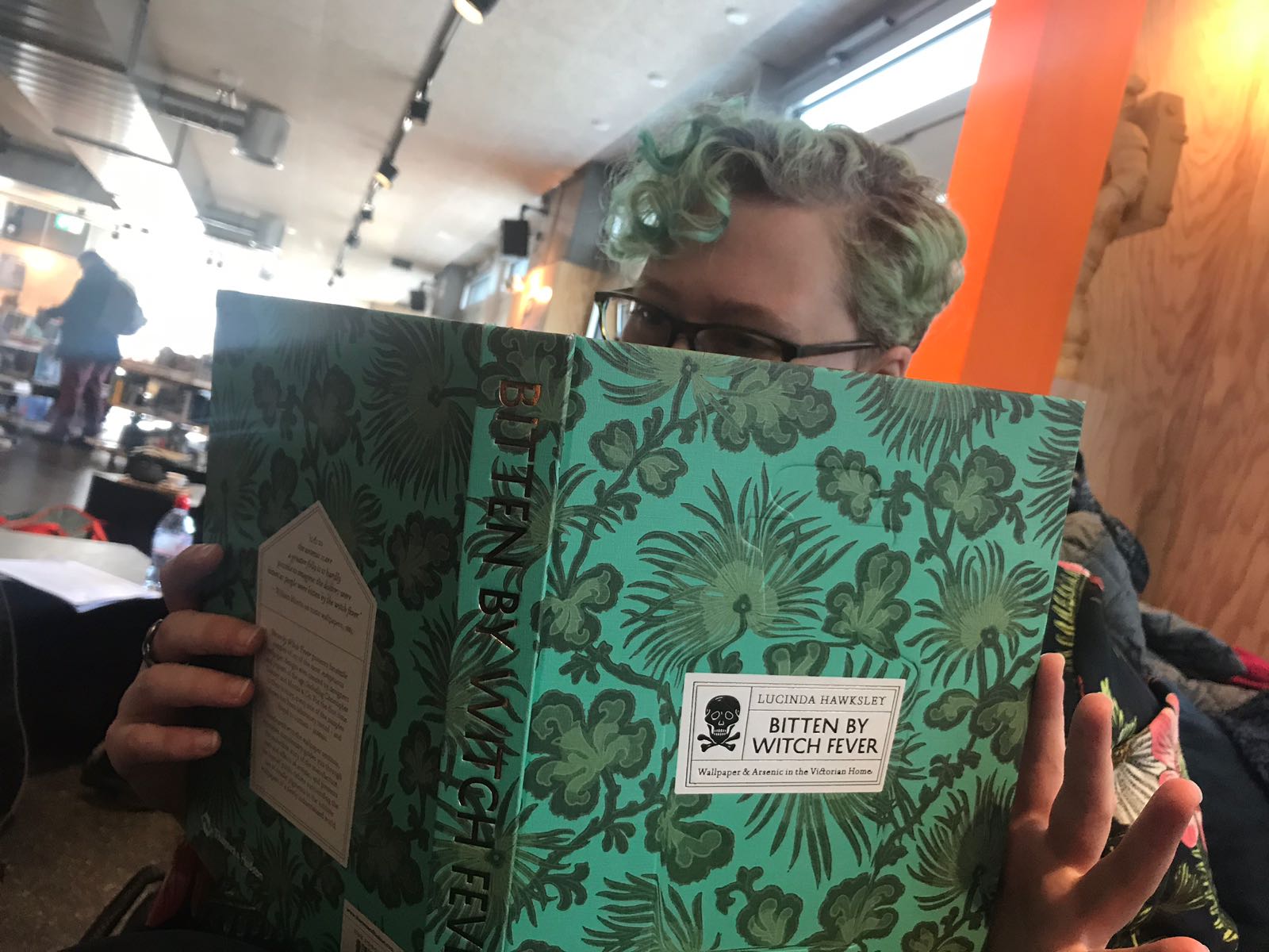 A person with curly green hair, reading a book with a green, flowery cover.